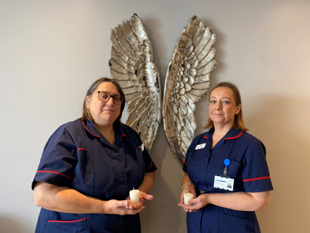 Two midwives stand in front of a wall that has an set of angel wings on. They both hold candles in their hands.