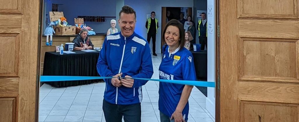 A man and woman dressed in Gillingham Football Club tops prepare to cut a blue ribbon