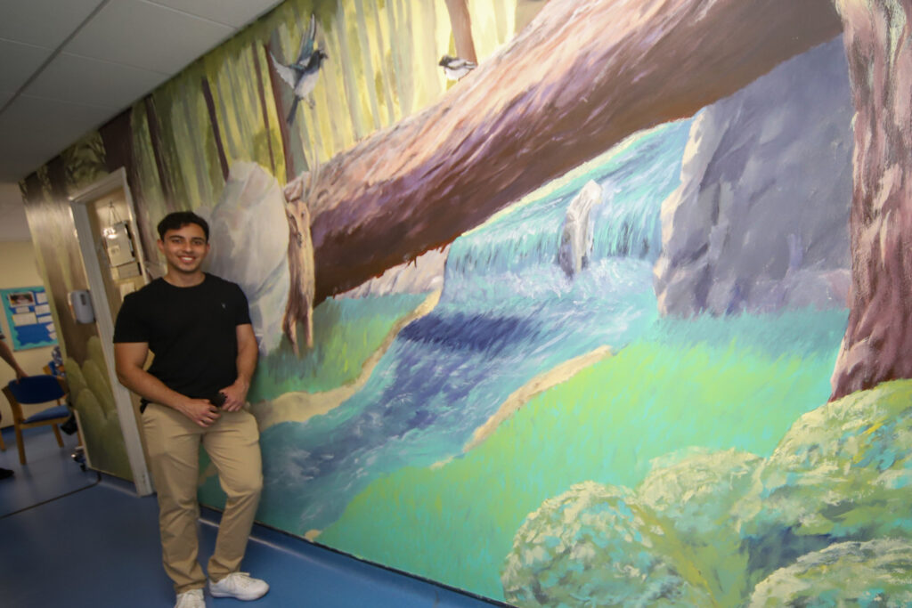 Mason Cheema stands next to the nature themed wall mural he created on the wall in Magpie Outpatients' Clinic