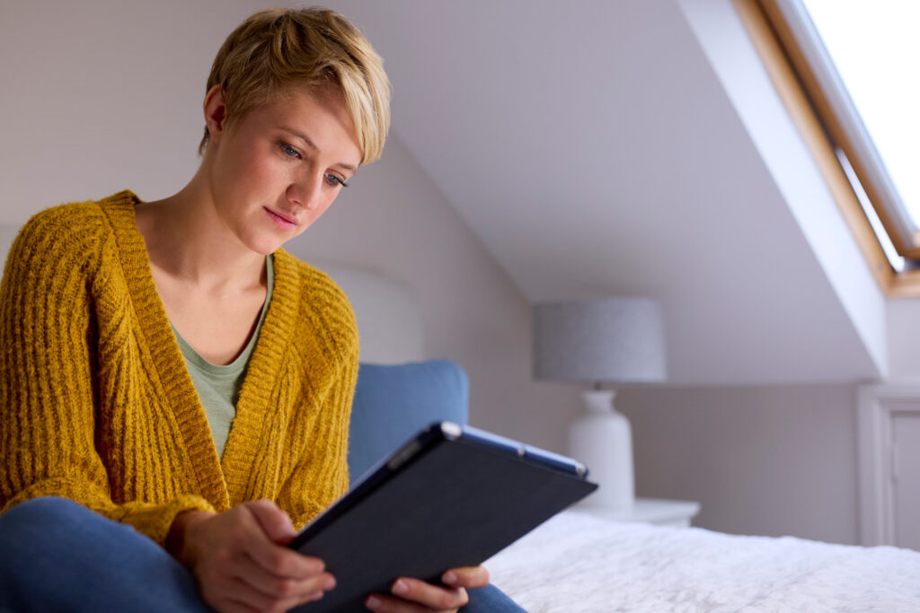Young woman sitting on a bed at home looking at digital tablet